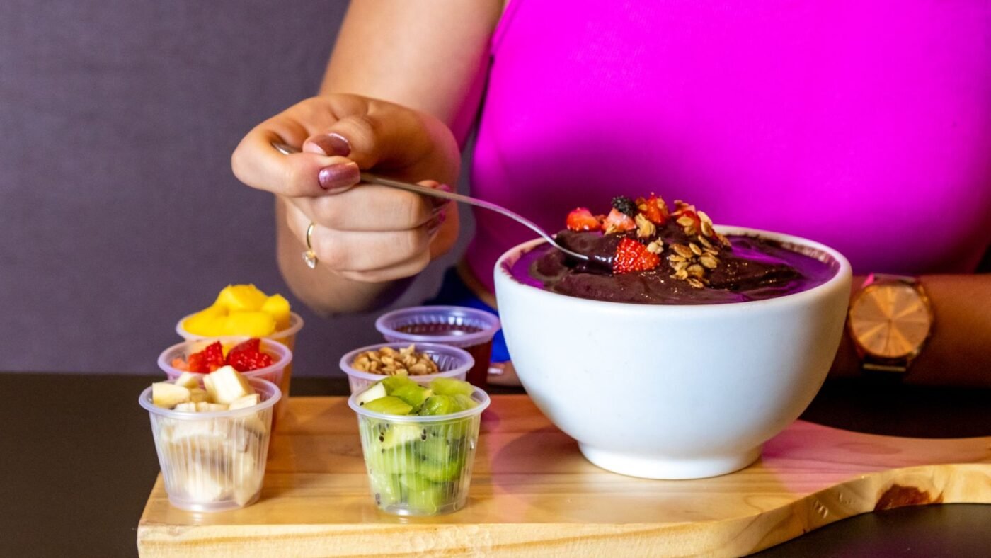 Tigela de açaí com granola e morango ao lado de potinhos de acompanhamentos como banana, manga e kiwi