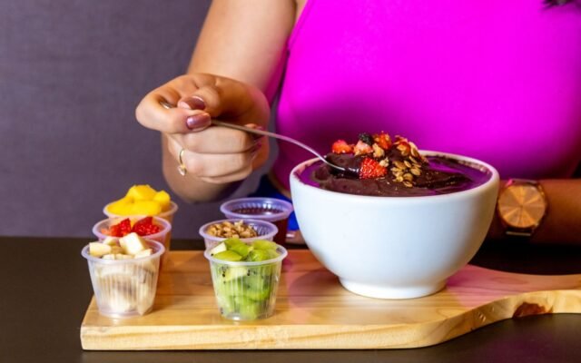 Tigela de açaí com granola e morango ao lado de potinhos de acompanhamentos como banana, manga e kiwi