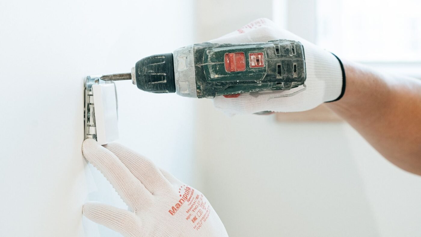Mãos de eletricista com luvas usando parafusadeira para instalar uma tomada na parede