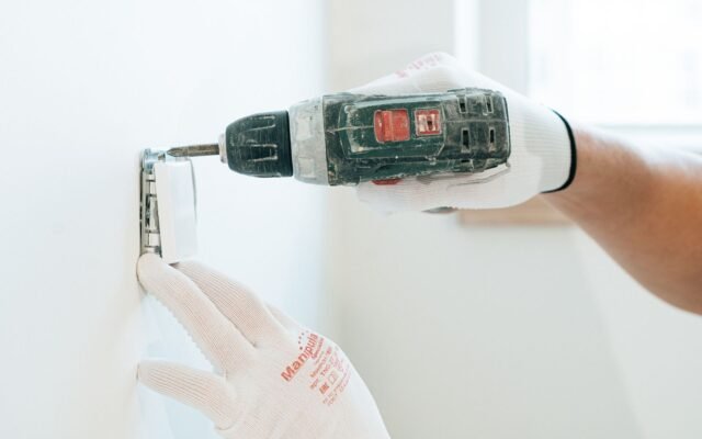 Mãos de eletricista com luvas usando parafusadeira para instalar uma tomada na parede