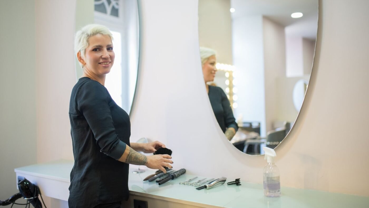 Cabeleireira sorrindo ao lado da bancada com espelho no salão de beleza