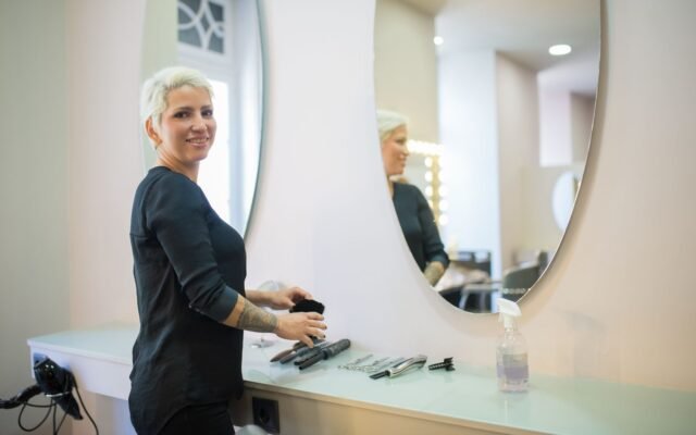 Cabeleireira sorrindo ao lado da bancada com espelho no salão de beleza