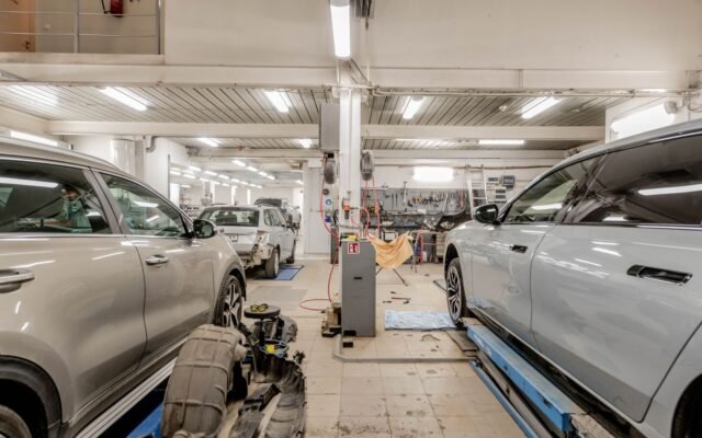 Oficina mecânica com vários carros em manutenção, um deles no elevador, e ferramentas na parede ao fundo