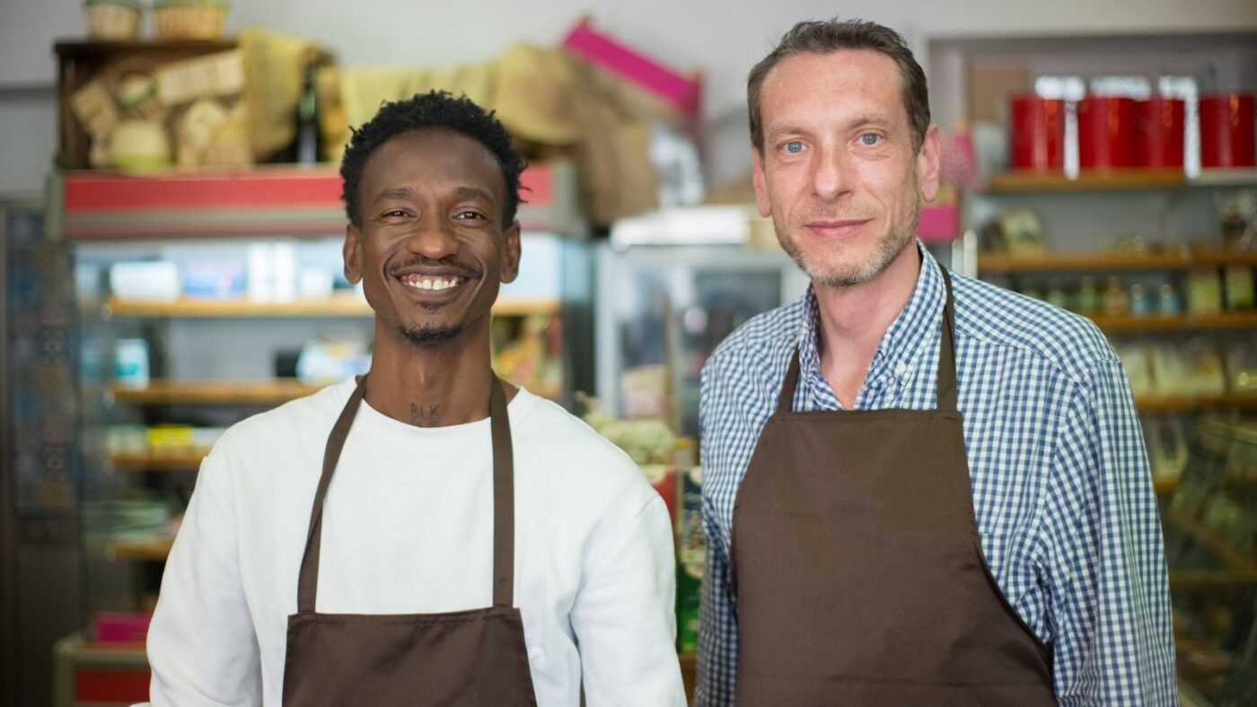 Dois homens de avental sorrindo lado a lado dentro de um pequeno mercado de bairro