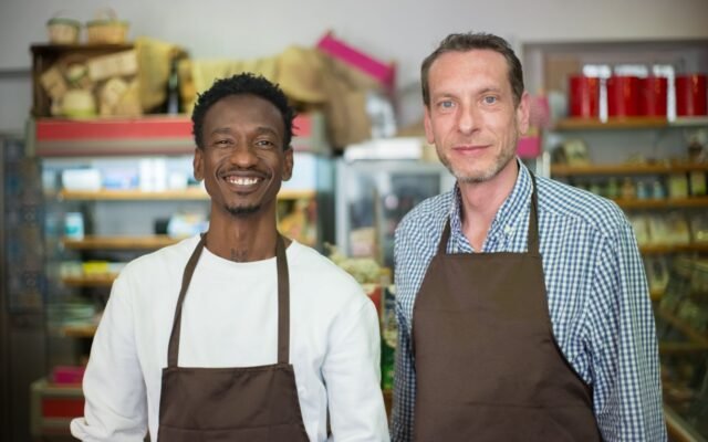 Dois homens de avental sorrindo lado a lado dentro de um pequeno mercado de bairro