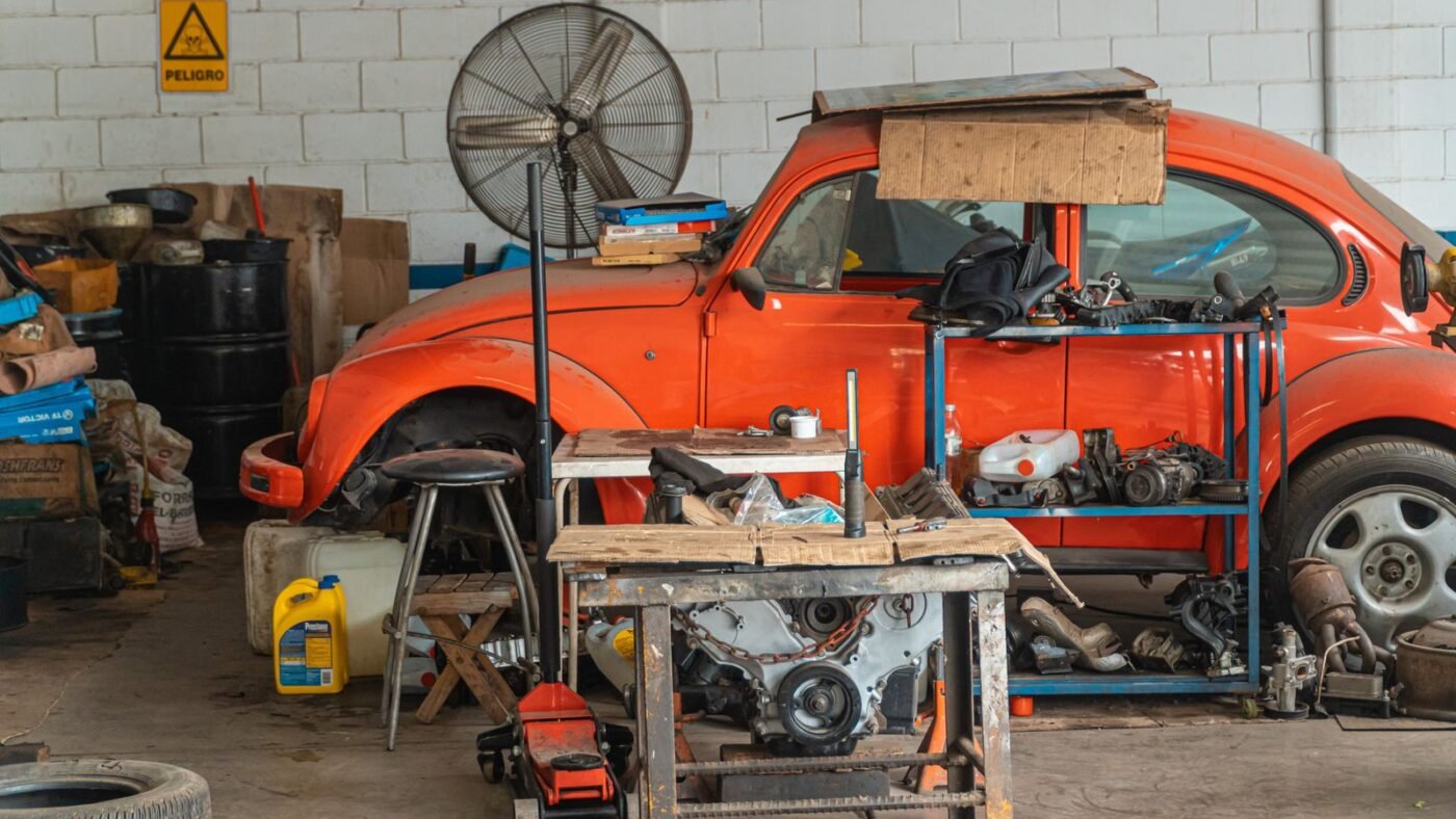 Oficina mecânica simples com Fusca laranja, bancada, macaco hidráulico, motor e peças espalhadas pelo chão