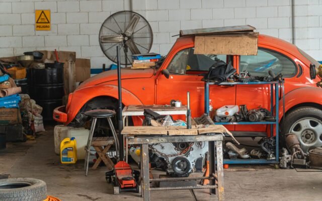 Oficina mecânica simples com Fusca laranja, bancada, macaco hidráulico, motor e peças espalhadas pelo chão