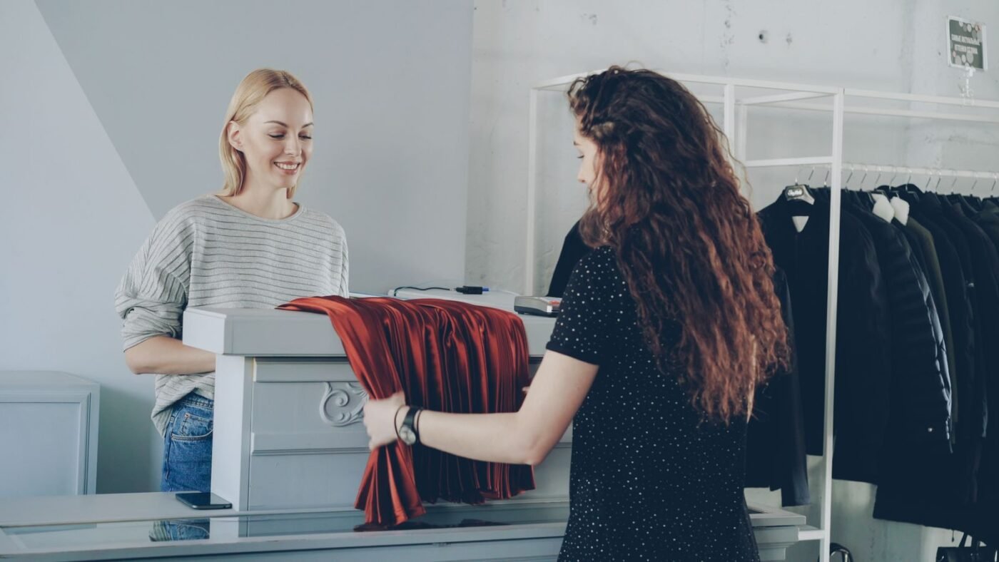 Atendente sorridente atrás do balcão de uma loja de roupas recebe uma peça vermelha de uma cliente, com arara de roupas ao lado