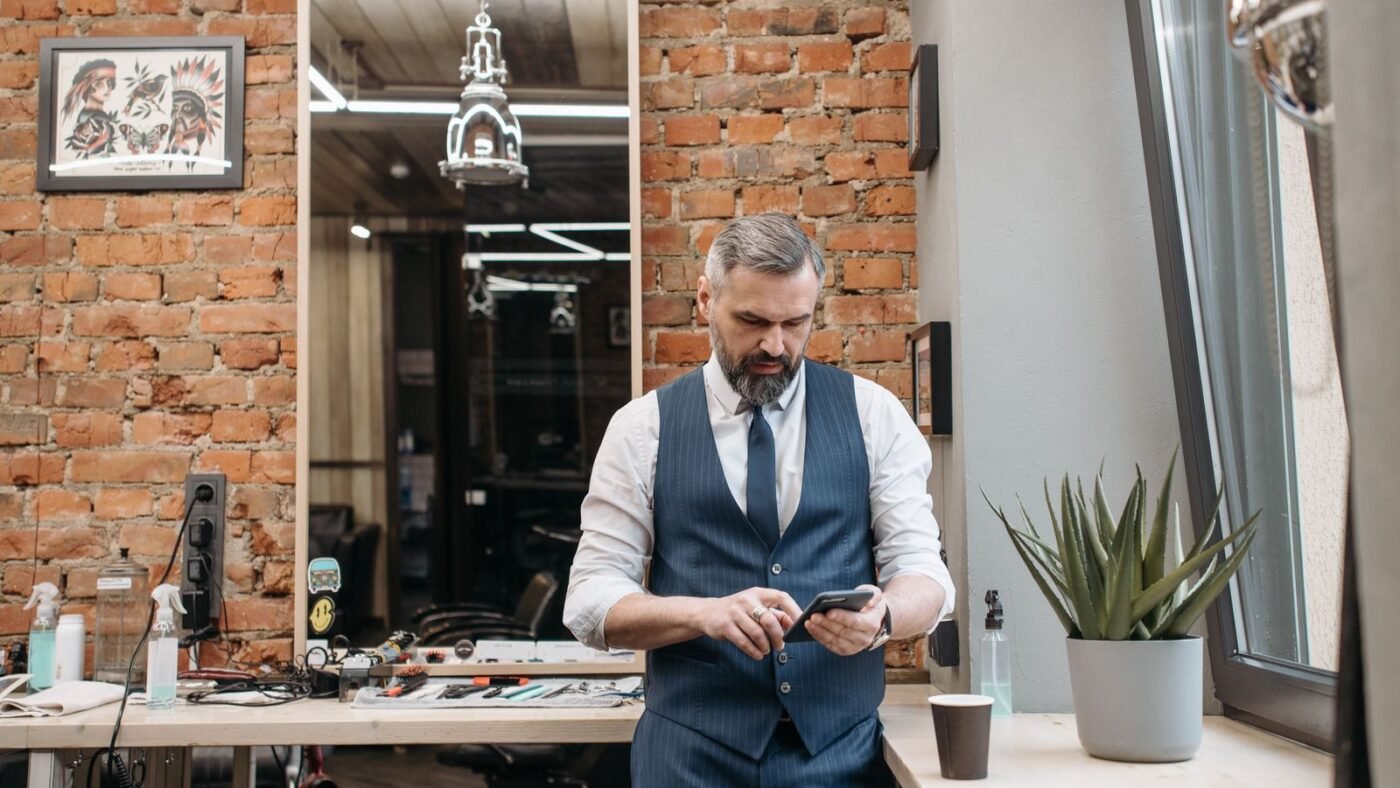 Dono de barbearia mexendo no celular encostado no balcão do salão