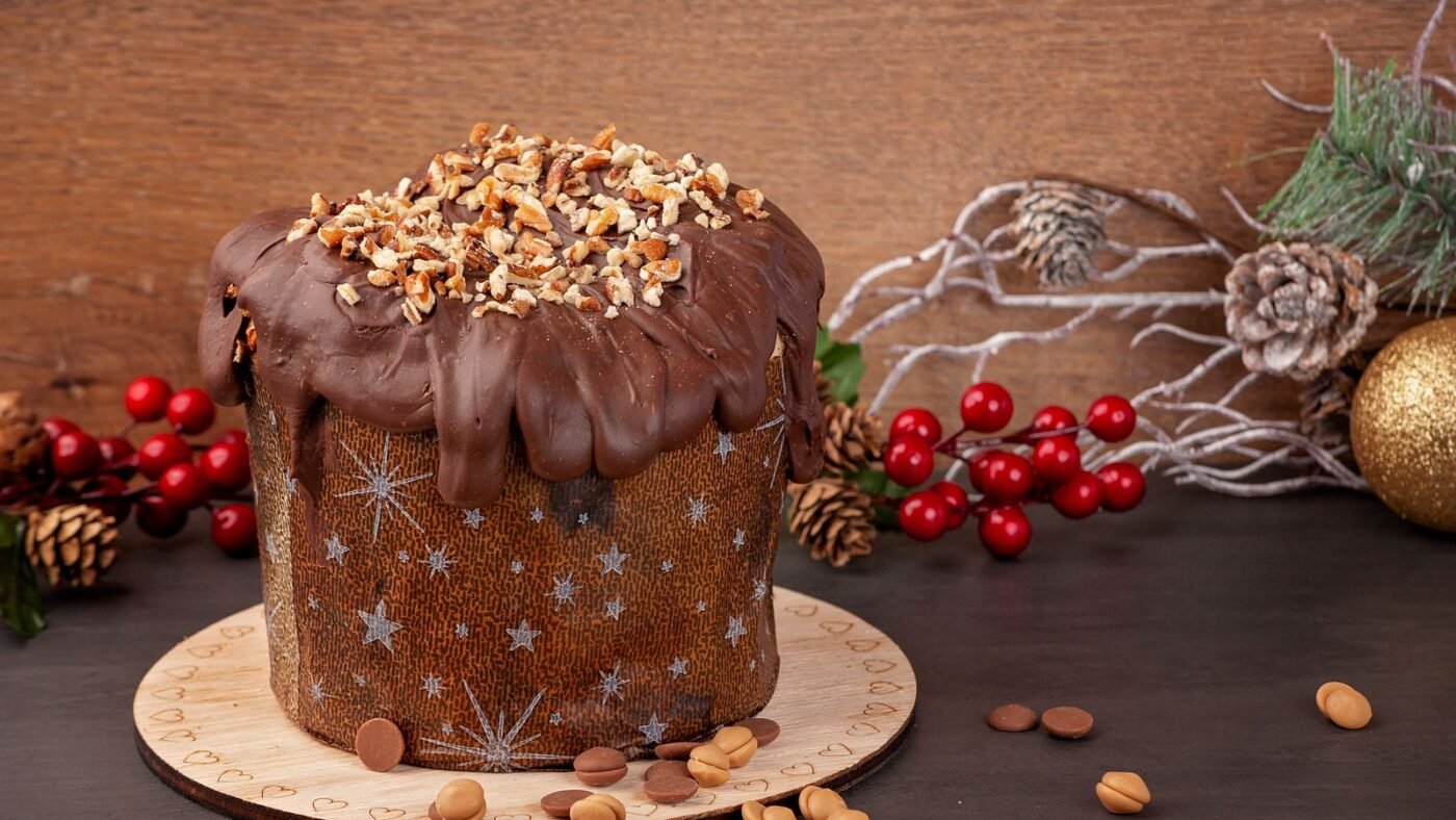 Panetone com cobertura de chocolate e castanhas picadas sobre tábua, com pinhas e enfeites de Natal ao fundo