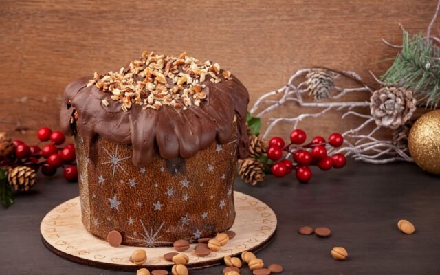 Panetone com cobertura de chocolate e castanhas picadas sobre tábua, com pinhas e enfeites de Natal ao fundo