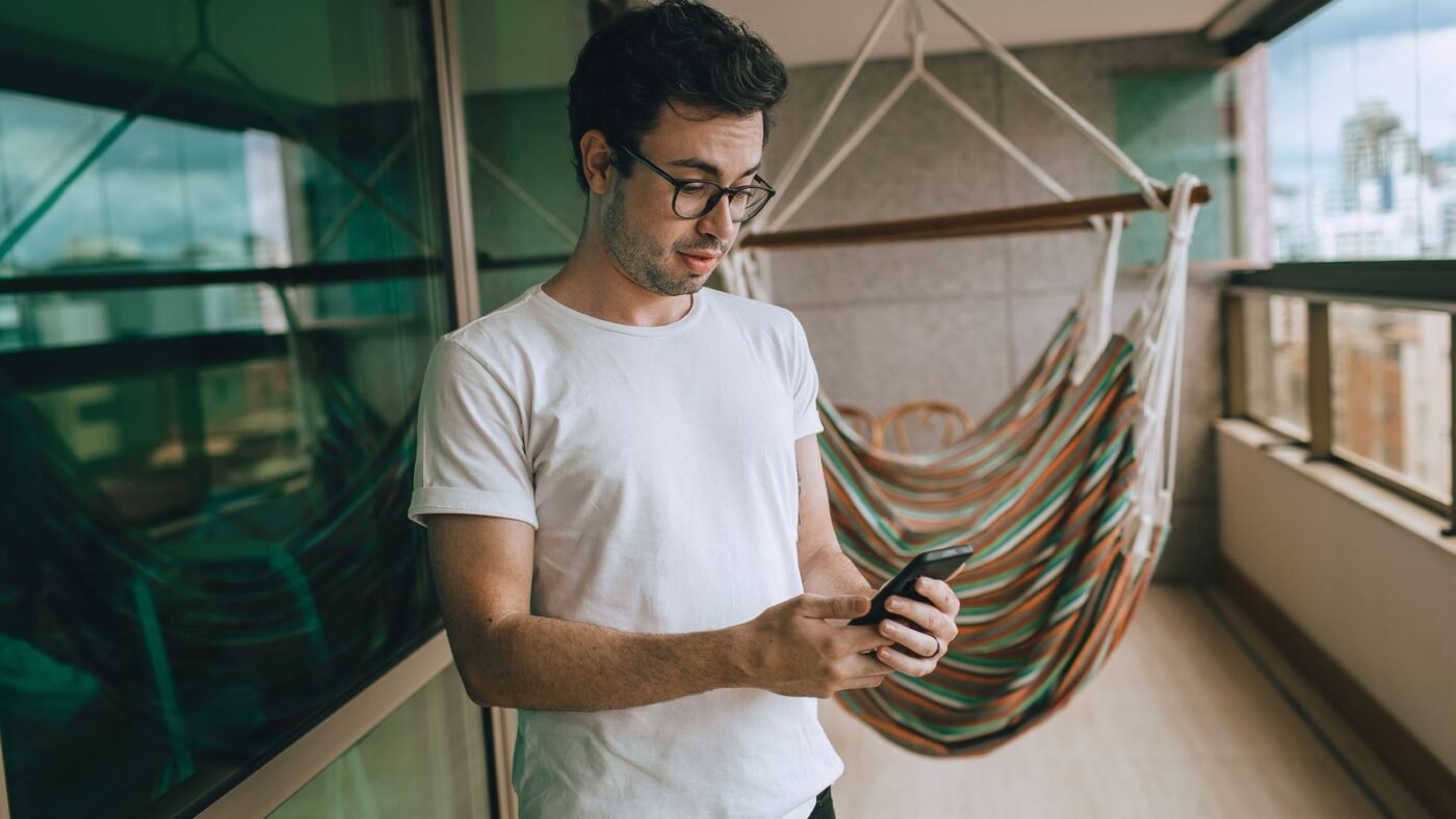 Homem de camiseta branca lendo uma mensagem no celular na varanda de casa