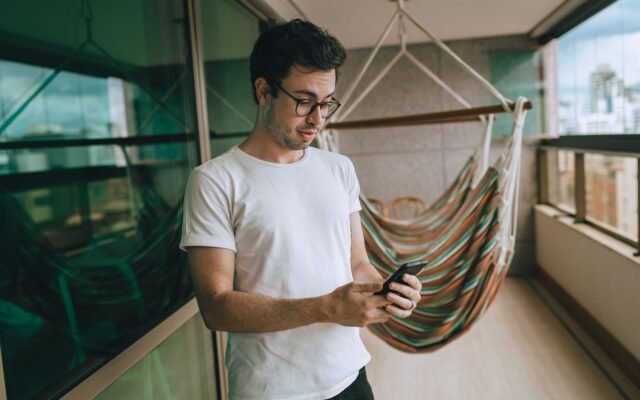 Homem de camiseta branca lendo uma mensagem no celular na varanda de casa