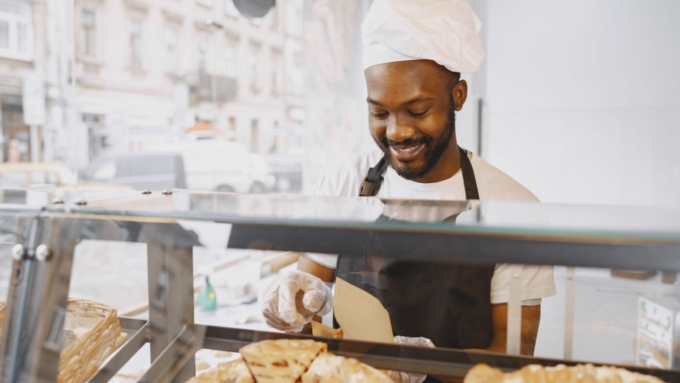 Funcionário de padaria de touca e avental sorrindo atrás da vitrine de tortas