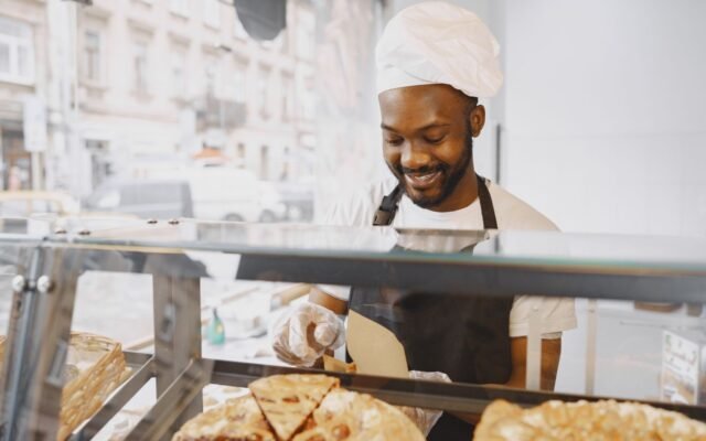 Funcionário de padaria de touca e avental sorrindo atrás da vitrine de tortas