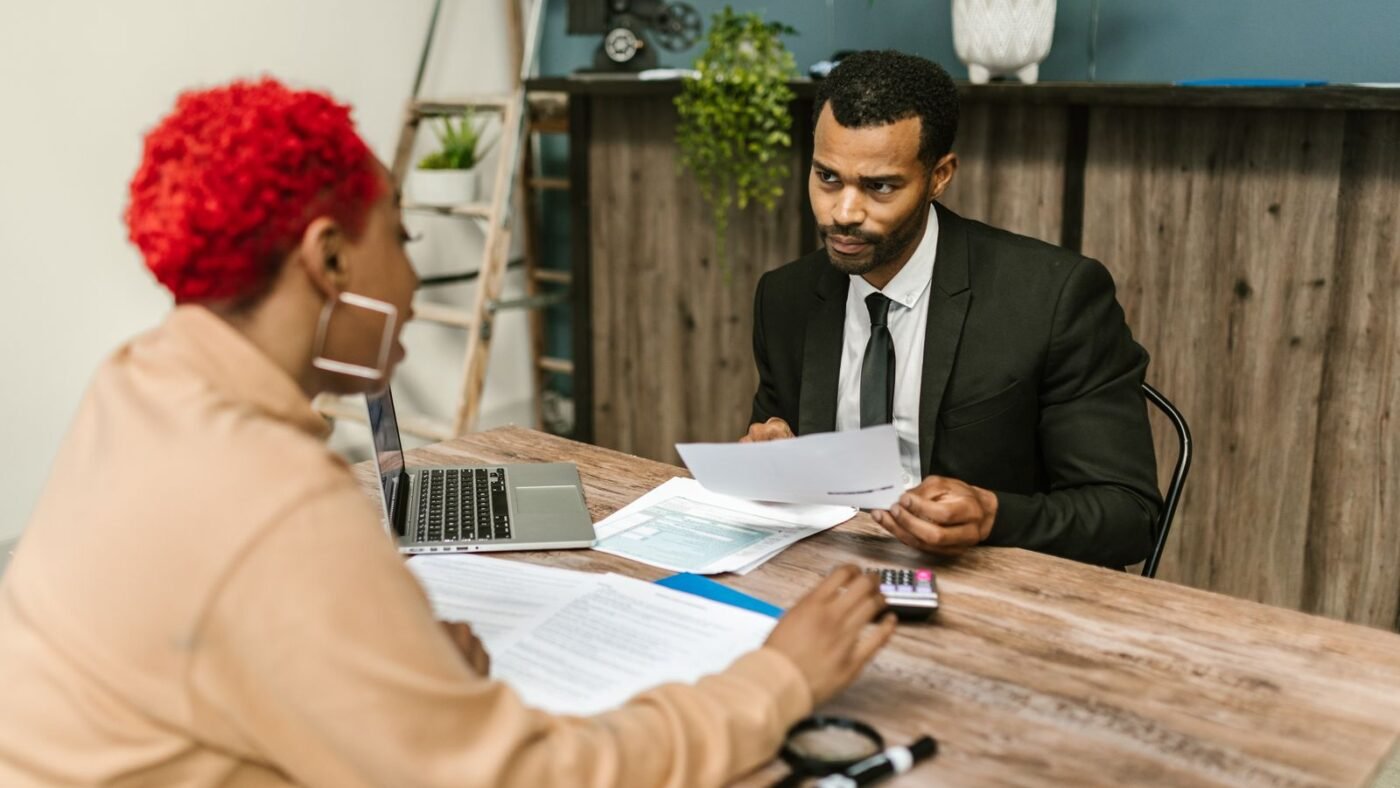 Homem de terno segura um papel e conversa com uma mulher de cabelo vermelho em uma mesa de madeira, com notebook, formulários e calculadora.