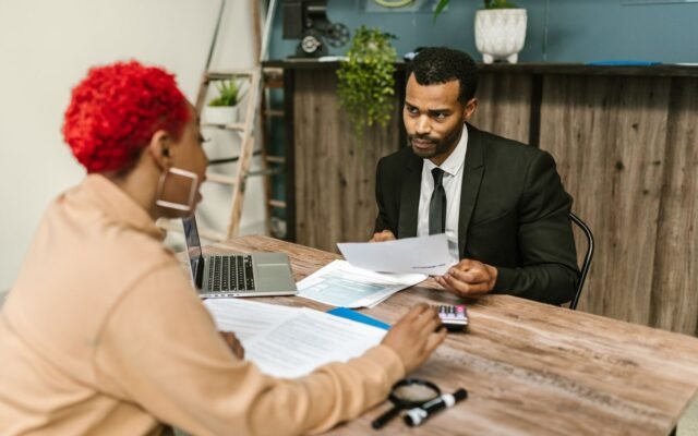 Homem de terno segura um papel e conversa com uma mulher de cabelo vermelho em uma mesa de madeira, com notebook, formulários e calculadora.