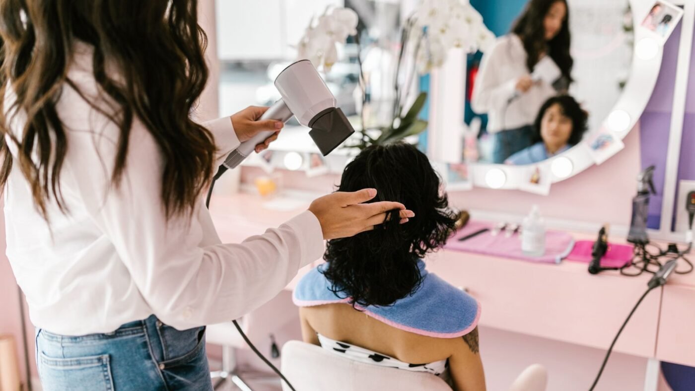 Cabeleireira secando o cabelo de uma cliente sentada diante do espelho no salão
