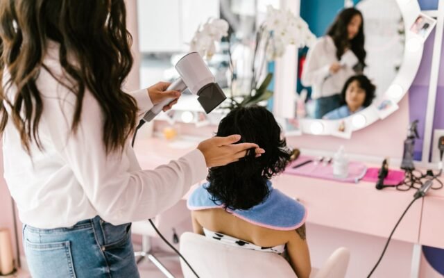 Cabeleireira secando o cabelo de uma cliente sentada diante do espelho no salão