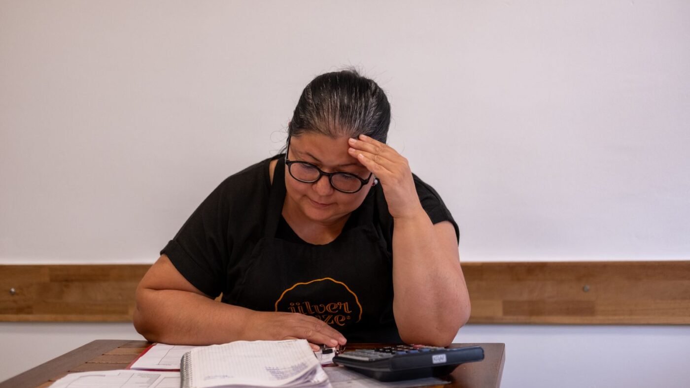 Mulher de óculos e avental preto, com a mão na testa, confere um caderno de contas e papéis ao lado de uma calculadora, em uma mesa de madeira.