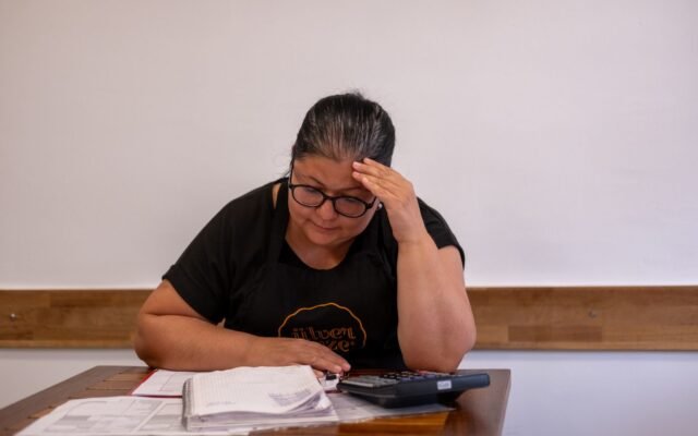 Mulher de óculos e avental preto, com a mão na testa, confere um caderno de contas e papéis ao lado de uma calculadora, em uma mesa de madeira.